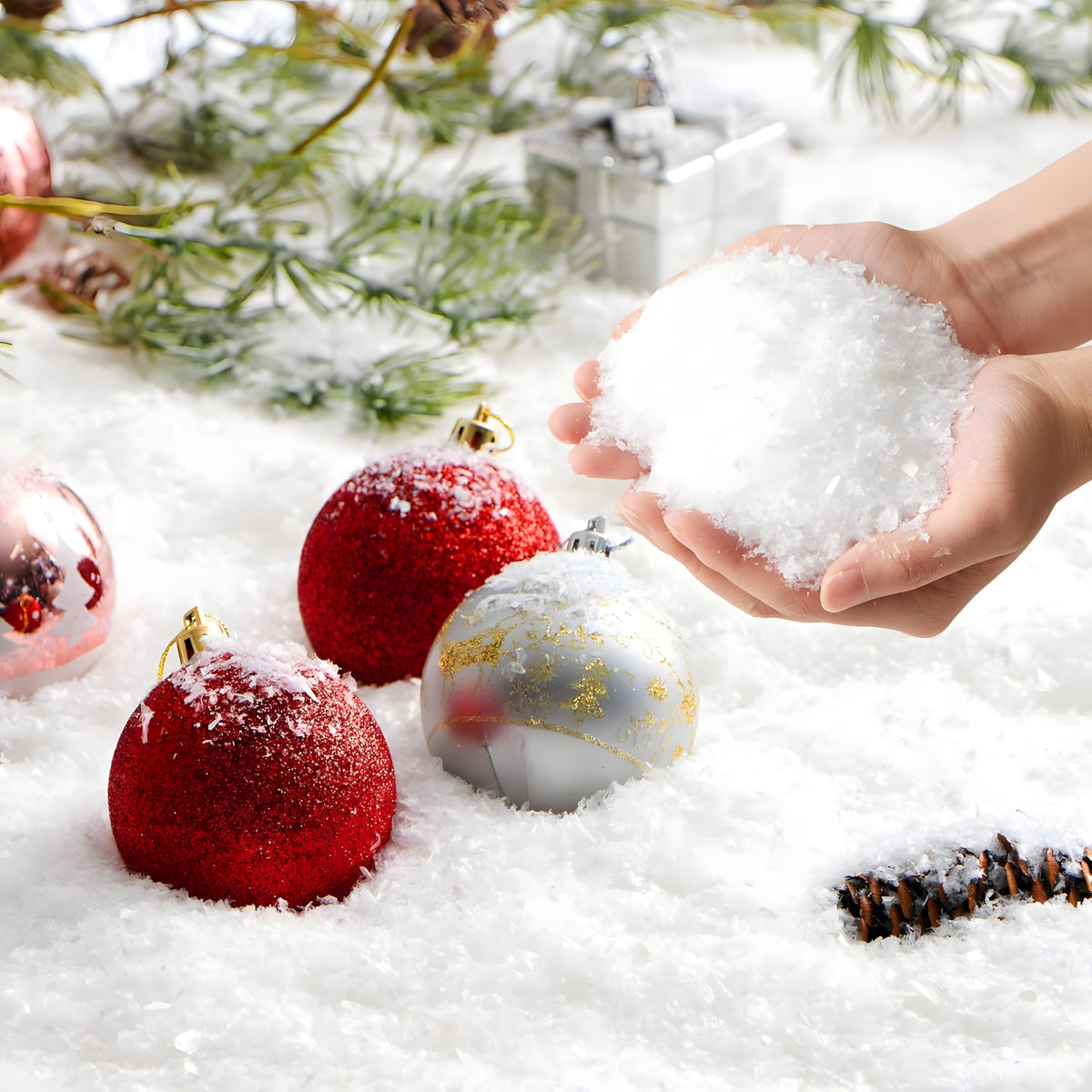 Two hands cupping a pile of white fluffy artificial snow over a winter background with red and silver Christmas tree baubles and pine branches